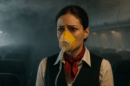 A concerned flight attendant wearing an oxygen mask in a dimly lit airplane cabin, with hazy smoke visible overhead.