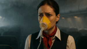 A concerned flight attendant wearing an oxygen mask in a dimly lit airplane cabin, with hazy smoke visible overhead.